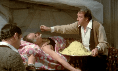 Σκηνή από την ιστορική ταινία του Μάρκο Φερέρι "Το Μεγάλο Φαγοπότι". Ο Ούγκο Τονιάτσι ταΐζει τον ήδη σκασμένο από το φαγητό Μισέλ Πικολί. Την πλάτη στον φακό έχει γυρισμένη ο Φιλίπ Νουαρέ.